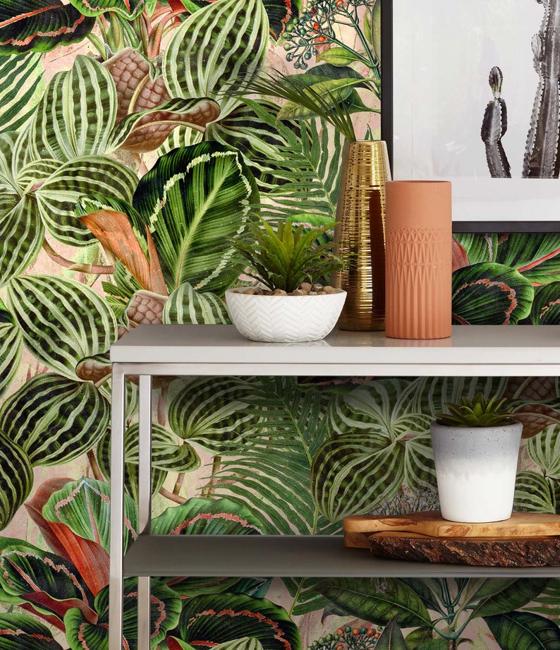May include: A white metal shelf unit with two shelves, featuring a tropical leaf patterned wallpaper backdrop. The top shelf has a white ceramic bowl with a small green plant, a gold metal vase, and a terracotta vase. The bottom shelf has a white ceramic pot with a small green plant and a wooden serving tray.
