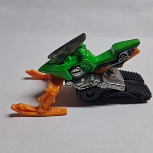 May include: A green and silver toy snowmobile with orange skis. The snowmobile has a black windshield and a black track. The number "02" is visible on the side. This toy is designed for play and collecting.