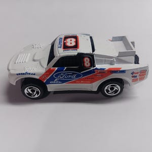 May include: White toy truck with blue and red accents. The truck has the number 8 on the roof and the word "Ford" on the side. The truck is a model of a Ford F-150.