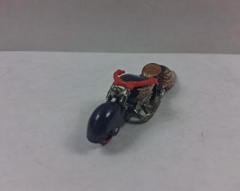 May include: A miniature motorcycle toy with a dark blue body, red handlebars, and silver-colored engine components. The seat and rear fender have a flame design. A small, circular emblem is on the rear.