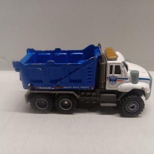 May include: A white and blue toy dump truck with a blue bed and a white cab. The truck has a "Tomica" logo on the side and says "Heavy Duty Truck". The truck has a white and blue "NYC Police" logo on the side of the cab.