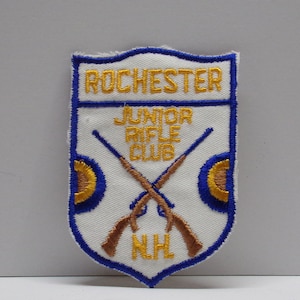 May include: White patch with blue border, featuring the text "Rochester Junior Rifle Club" and "NHL" in gold embroidery. Two crossed rifles are depicted in brown embroidery.