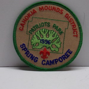 May include: A tan and green embroidered patch with the text "Cahokia Mounds District Patriots Park 1996 Spring Camporee".