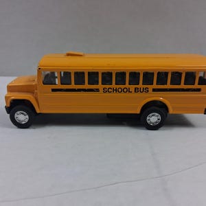 May include: A yellow toy school bus with black wheels and the words "SCHOOL BUS" printed in black on the side. The bus has multiple windows and a roof-mounted light. The toy is set against a neutral background.