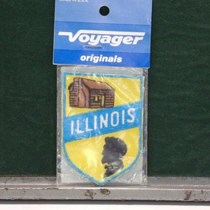 May include: A yellow and blue embroidered patch with the word "ILLINOIS" and a small cabin and a silhouette of a person. The patch is packaged in a clear plastic bag with a Voyager Originals header.