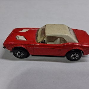 May include: A red toy car with a white convertible top. The toy car has black wheels and white accents on the hood. The car is a vintage model, likely from the 1970s.