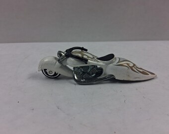 May include: A white toy motorcycle with gold flame designs on the sides. The motorcycle has a black seat and handlebars, and chrome-colored engine components. The toy is a miniature replica of a custom chopper-style motorcycle.