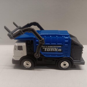 May include: A blue and white Tonka City Sanitation garbage truck toy. The truck features a blue cab and waste container, with the words "City Sanitation" and "Tonka" printed on the side. The truck has black wheels and a gray lifting mechanism.