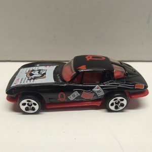 May include: Black toy car with red accents and playing card designs. The car has a Queen of Hearts design on the roof and a Jack of Hearts design on the side. The car is a 1967 Chevrolet Corvette.