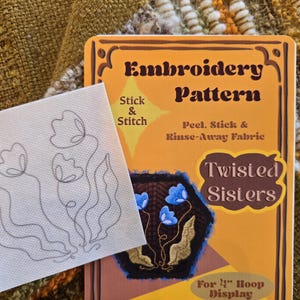 May include: An embroidery pattern kit with the text "Embroidery Pattern" and "Twisted Sisters". The kit includes a peel, stick, and rinse-away fabric. The design features blue flowers. The kit is for a 4" hoop display.