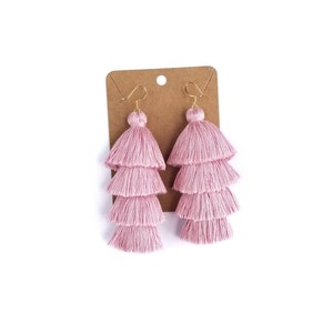 May include: A pair of light pink tiered tassel earrings featuring four stacked layers of soft thread fringe. Each earring is attached to a gold-toned metal hook and is displayed on a brown rectangular cardboard jewellery backing card.