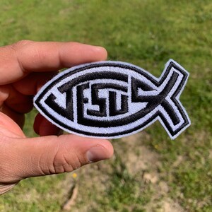 May include: A white and black embroidered iron-on patch in the shape of a fish with the word "JESUS" spelled out inside the fish.