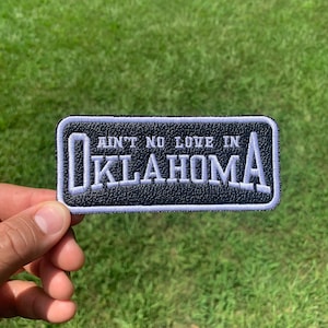 May include: Black leather patch with white stitching. The patch says "Ain't No Love In Oklahoma" in white lettering.