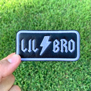 May include: A black leather patch with white stitching and a white lightning bolt design. The text "Lil Bro" is embroidered in white on the patch.