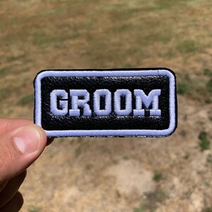 May include: A black leather patch with a white embroidered border and the word "GROOM" in white lettering.