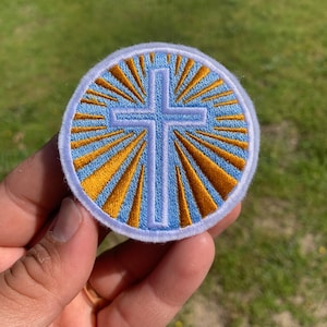 May include: A round, blue and gold embroidered patch with a white cross in the center. The cross is surrounded by gold rays radiating outwards.
