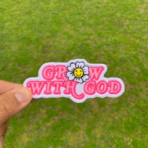 May include: Pink and white embroidered patch with the text "GROW WITH GOD" and a white flower with a yellow smiley face in the center.
