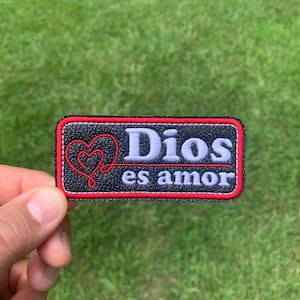 May include: Black leather patch with red stitching and a white embroidered heart. The text "Dios es amor" is embroidered in white.