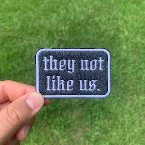 May include: Black leather patch with white stitching. The patch says "they not like us."