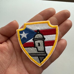 May include: A yellow, red, white, and blue embroidered patch in the shape of a shield with a fort and the Puerto Rican flag.