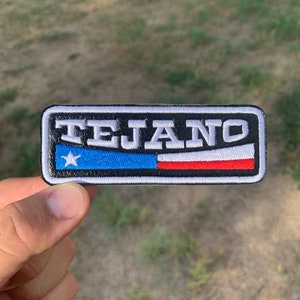 May include: A black and white embroidered patch with the word "TEJANO" in white lettering. The patch features a blue and red stripe with a white star in the upper left corner.