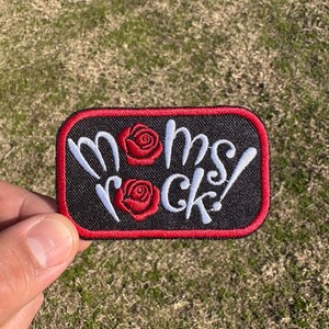 May include: Black iron-on patch with red stitching and white text that reads "Moms Rock!" with two red roses.