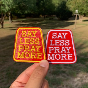 May include: Two red fabric patches with yellow and white stitching. The patches are shaped like a trapezoid and have the text "SAY LESS PRAY MORE" on them.