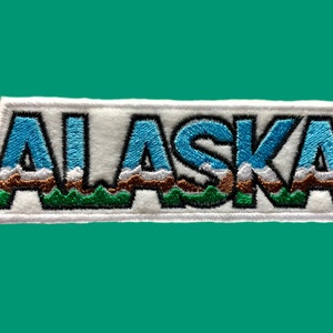 May include: White embroidered patch with the word "ALASKA" in blue, green, and brown. The letters are outlined in black and have a mountain range design.