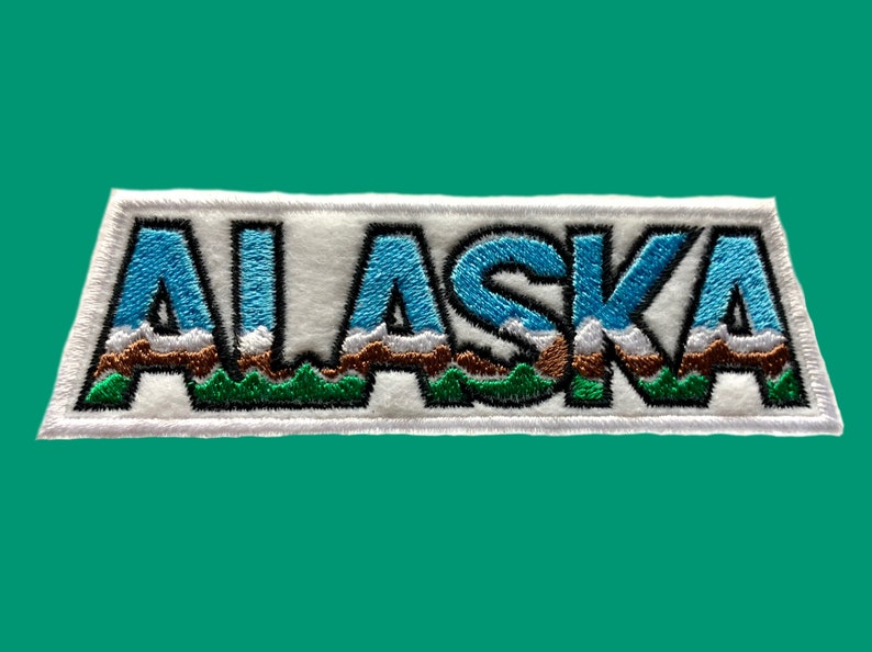 May include: White embroidered patch with the word "ALASKA" in blue, green, and brown. The letters are outlined in black and have a mountain range design.