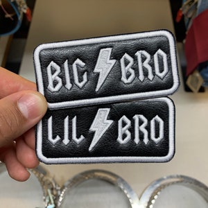 May include: Two black leather patches with white stitching. The top patch says "BIG BRO" with a lightning bolt in between the words. The bottom patch says "LIL BRO" with a lightning bolt in between the words.