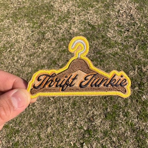May include: A brown and yellow embroidered patch shaped like a hanger with the text "Thrift Junkie" in black.