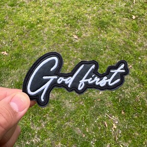 May include: Black and white embroidered patch with the text "God first" in a cursive font.