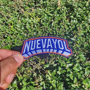 May include: A blue and orange embroidered patch with the word "NUEVAYOL" in white block letters. The patch has a curved shape and is held against a background of green foliage.
