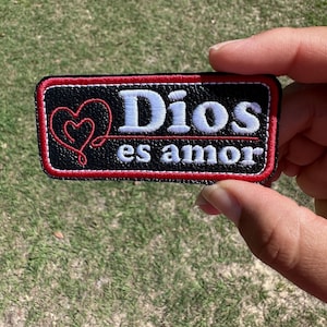 May include: Black leather patch with red stitching and white embroidery. The patch features a heart design and the text "Dios es amor".