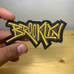 May include: A black and gold embroidered patch with the word "BROOKLYN" in a stylized font.