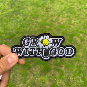 May include: A black and white embroidered patch with a white daisy with a yellow smiley face in the center. The text "GROW WITH GOD" is in white with a black outline.