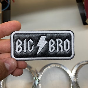 May include: Black leather patch with white stitching. The patch features a white lightning bolt and the text "BIG BRO" in white.