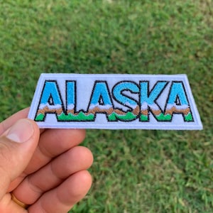 May include: White embroidered patch with the word "ALASKA" in blue letters with a brown and green mountain range behind the letters.