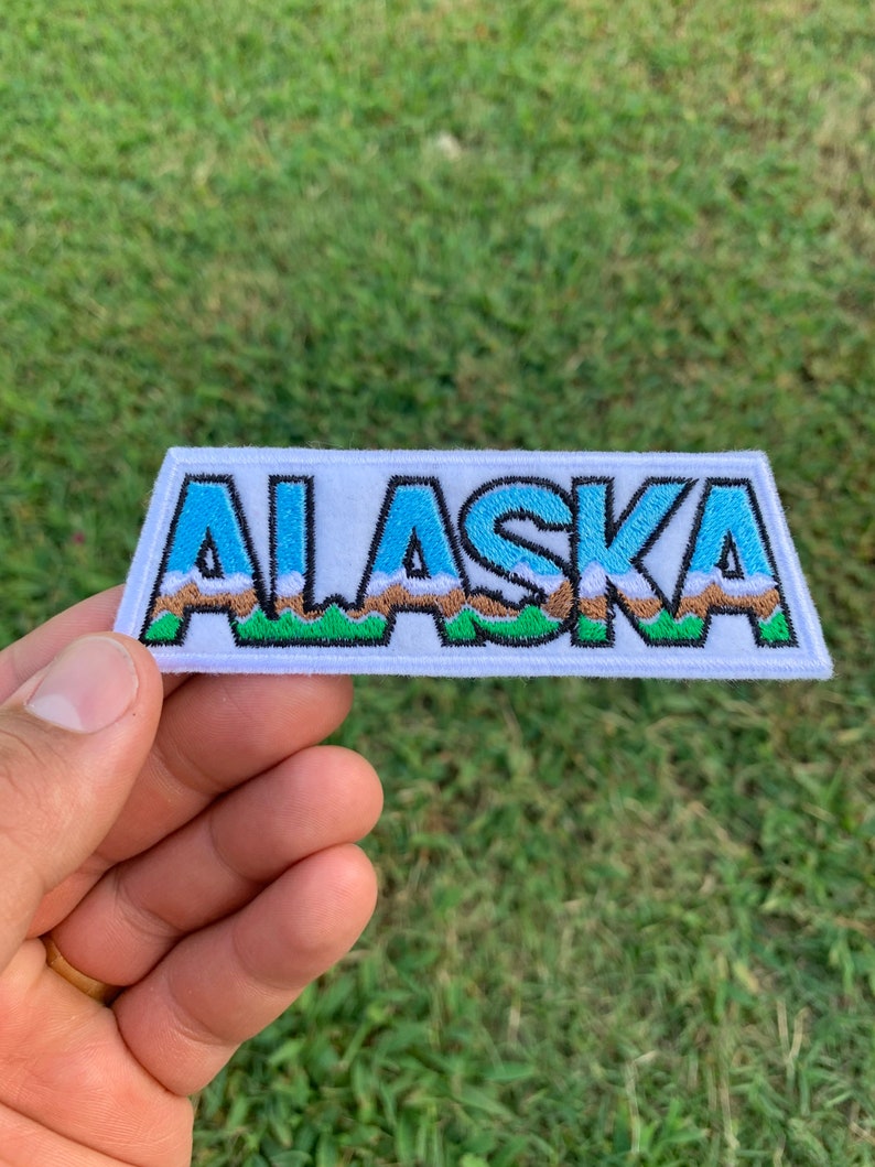 May include: White embroidered patch with the word "ALASKA" in blue letters with a brown and green mountain range behind the letters.