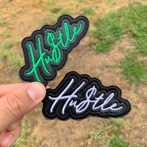May include: Two black fabric patches with the word "Hustle" embroidered in green and white thread.