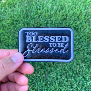May include: A black leather patch with white stitching. The patch says "Too Blessed To Be Stressed".