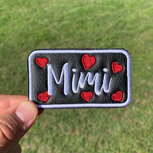 May include: Black leather patch with white stitching and the name "Mimi" embroidered in white. The patch is decorated with red hearts.