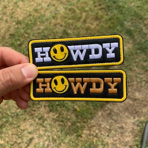 May include: Two black fabric patches with yellow borders. Both patches have a smiley face and the word "HOWDY" in white and gold lettering.