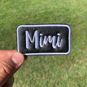 May include: A black leather patch with a white border and the name "Mimi" embroidered in white.