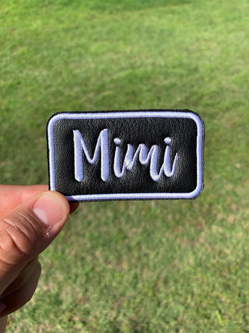 May include: A black leather patch with a white border and the name "Mimi" embroidered in white.