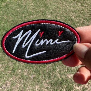May include: A black leather patch with a white embroidered cursive word "Mime" and two red hearts. The patch has a red stitched border.