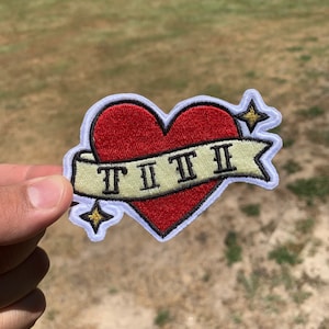 May include: A red heart with a white banner that says "TIII" in Roman numerals. The heart has a white outline and two gold stars on either side.