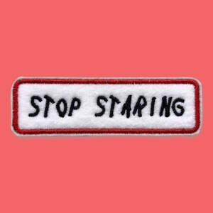 May include: White embroidered patch with a red border. The patch says "STOP STARING" in black lettering.