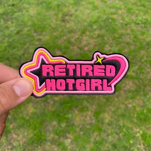 May include: A pink and yellow embroidered patch with the text "Retired Hotgirl" in black lettering.