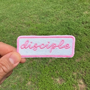 May include: A rectangular patch with the word "disciple" embroidered in pink cursive script on a white background. The patch is outlined with a pink border. The patch is held against a background of green grass.